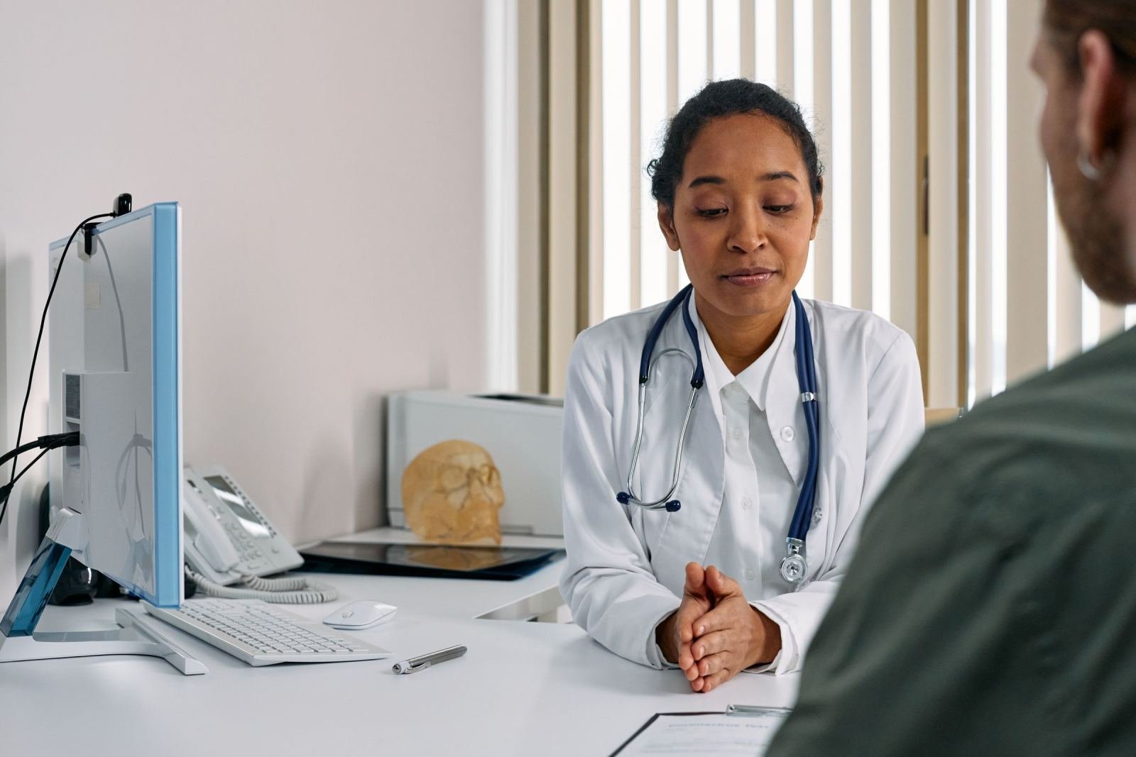 A clinician in a white coat and stethoscope talks with a patient in an office