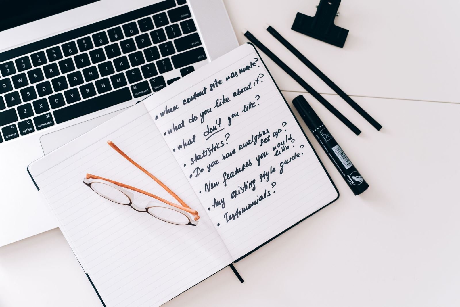A notebook with a handwritten list of questions next to a laptop and glasses