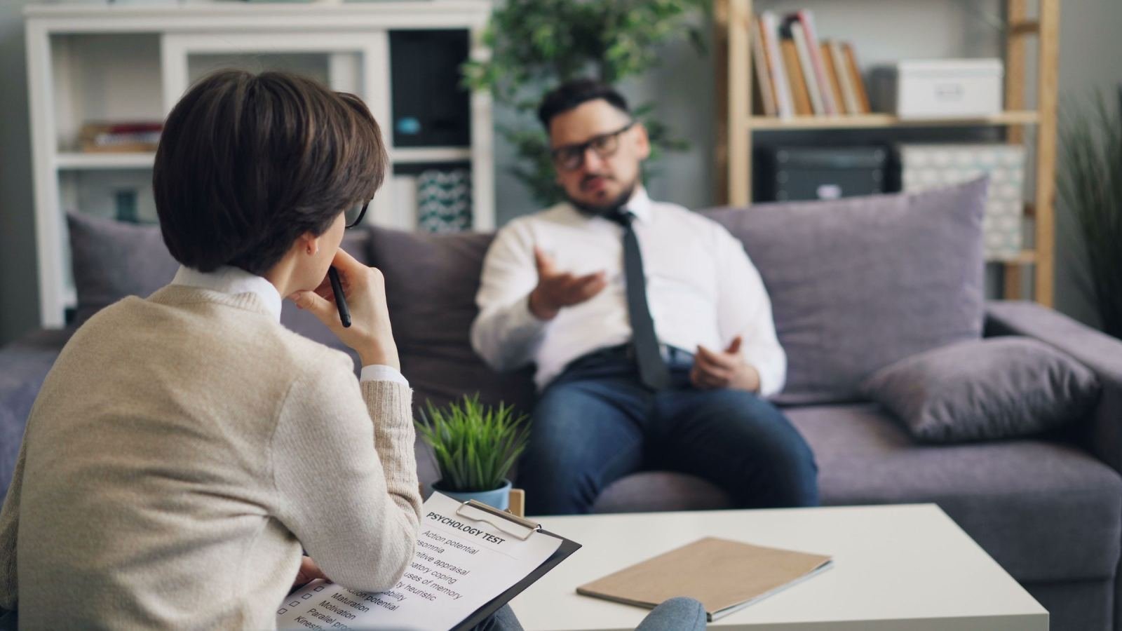 In a bright office, a clinician with a clipboard talks with a man wearing a white shirt.