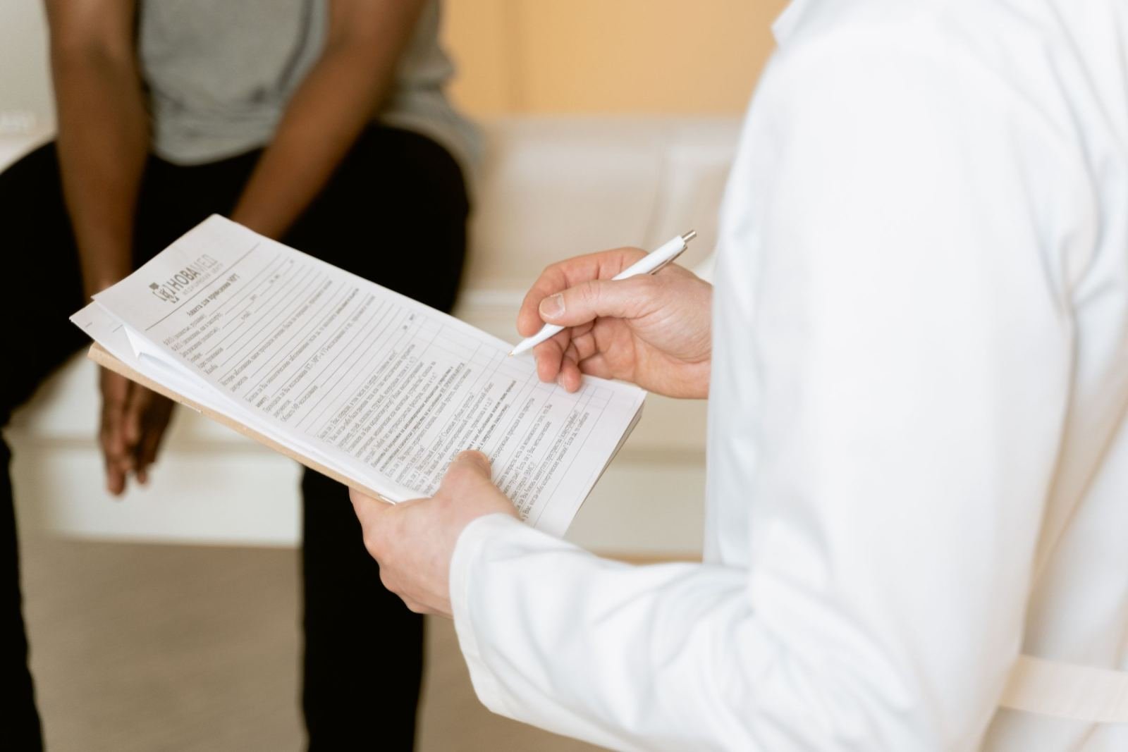A clinician in a white coat writes on a clipboard while a patient sits in the background