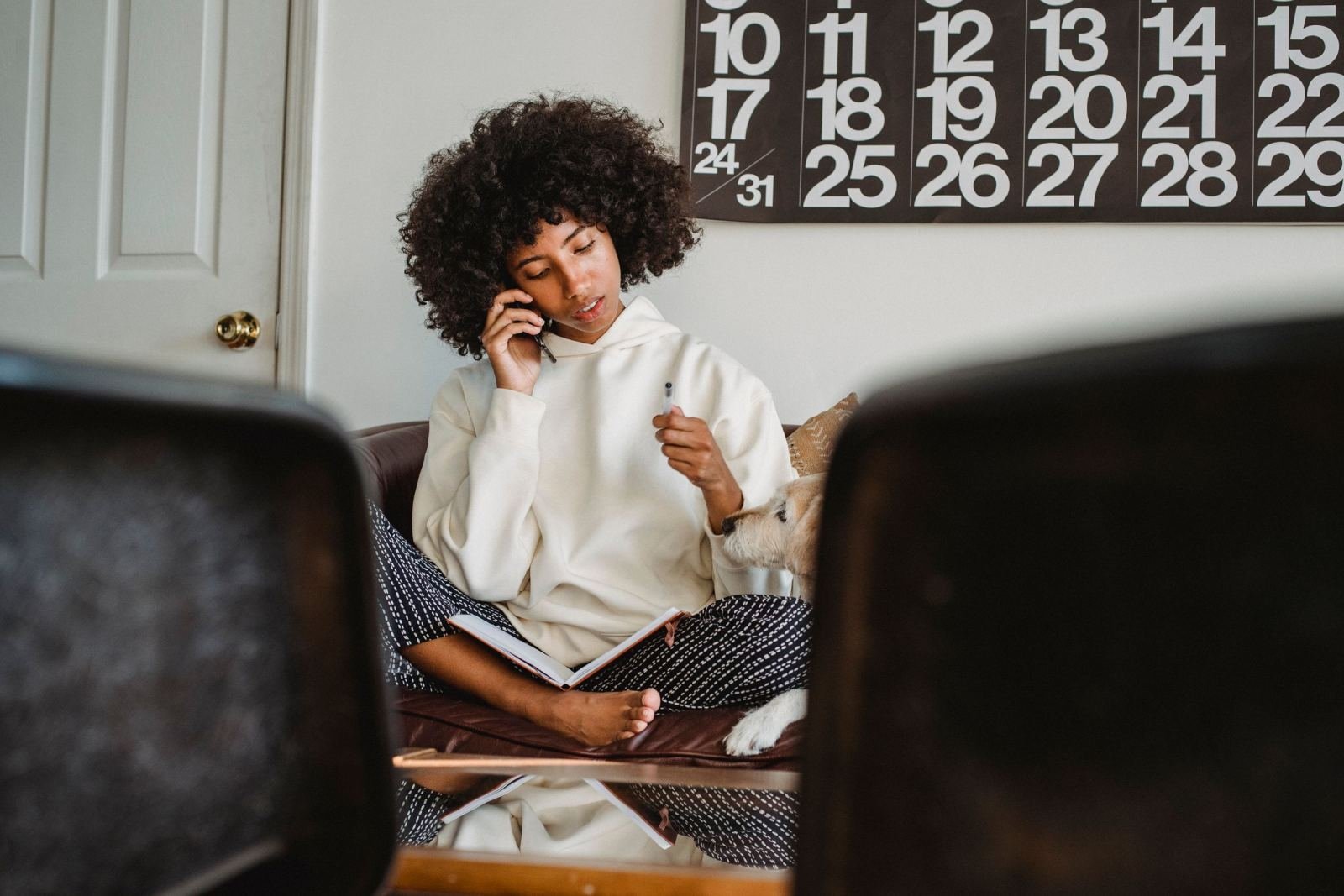 A woman sits on a couch with a dog. She holds a phone and writes notes in a pad.