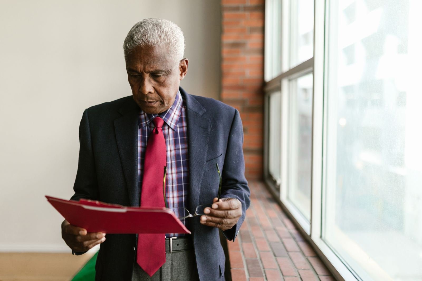 An older man in a suit reviews documents in a red folder by a window