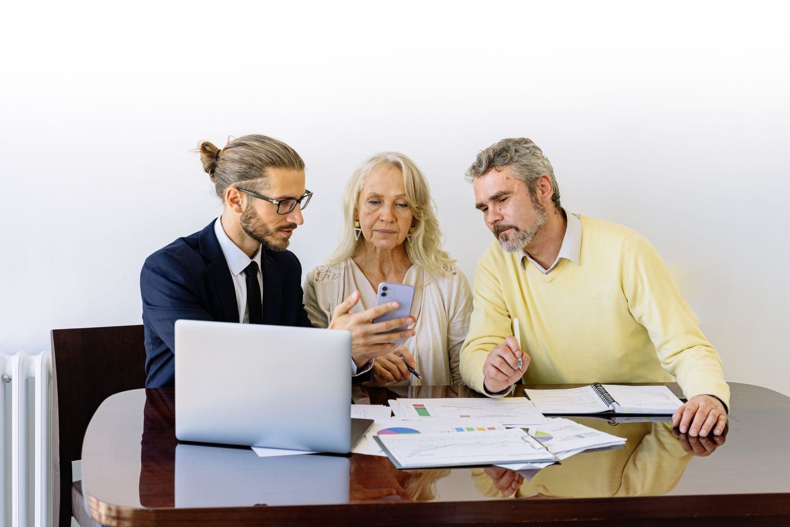Three people look over a phone, a laptop, and paper charts while seated at a table.