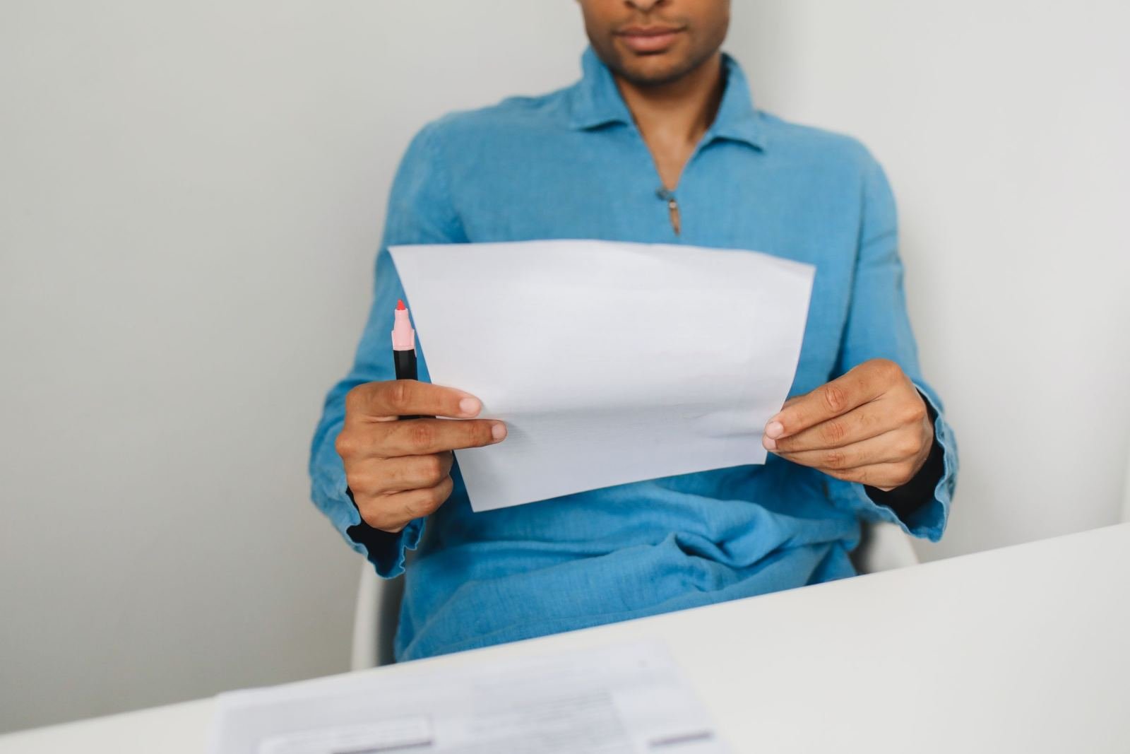 Someone in a blue shirt sits at a white table holding a paper and a red pen.