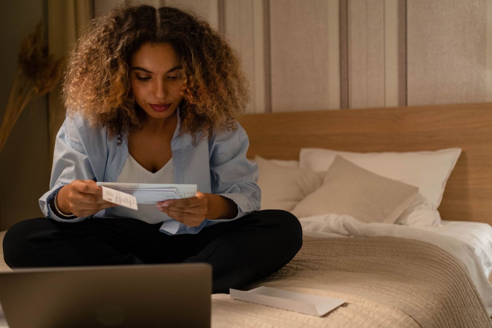 A woman sits on her bed reading a letter with a laptop open in front of her