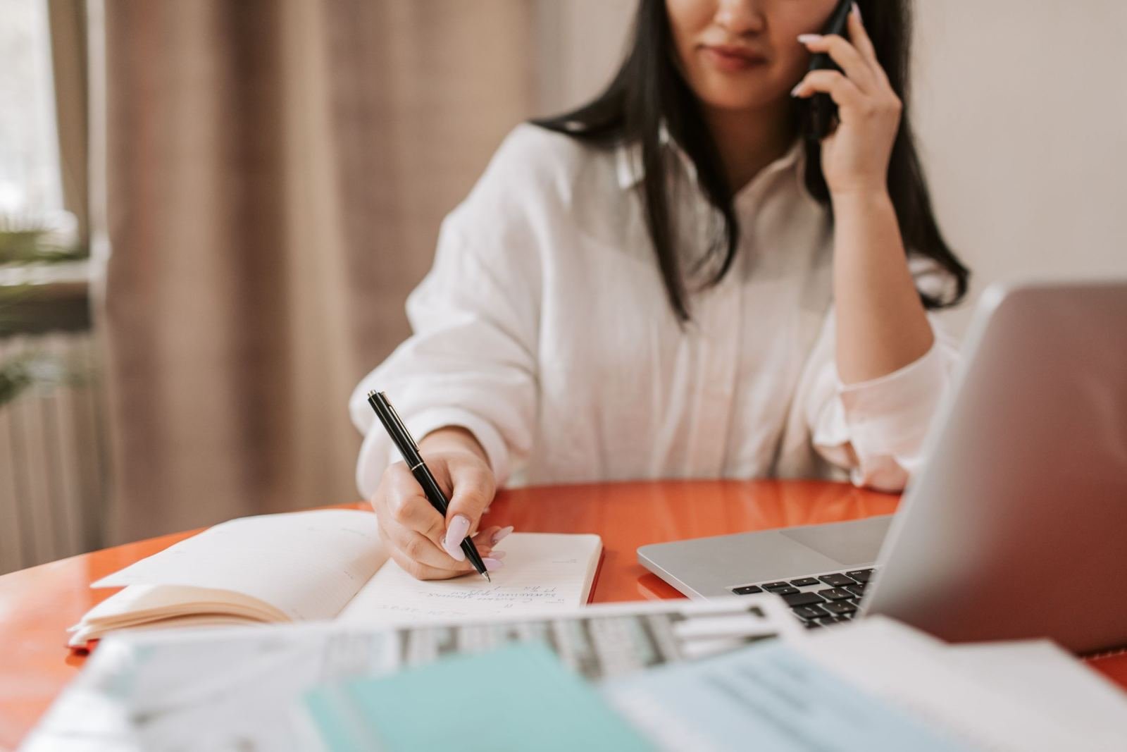 A woman talks on the phone while writing in a notebook at a desk with a laptop
