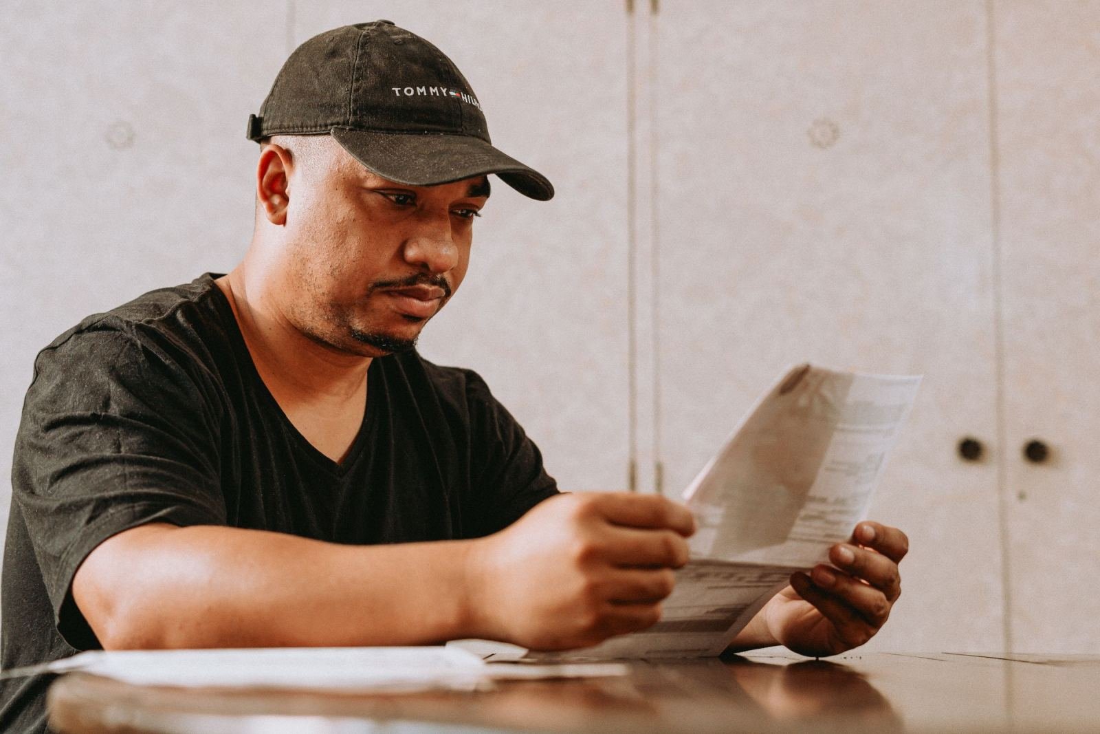 A man wearing a black cap sits at a table and reads a paper.