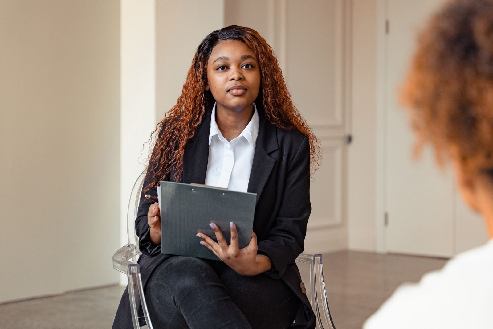 A professional woman holds a clipboard while seated in a clear chair at a meeting.