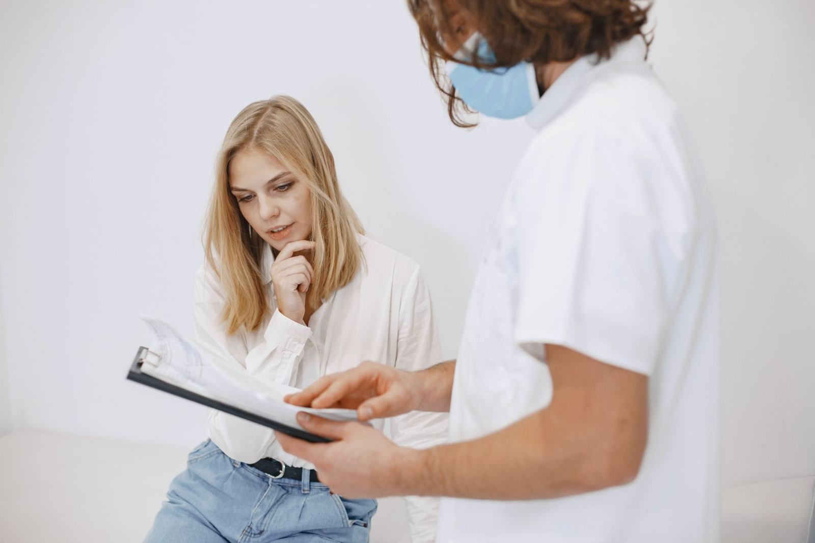 A doctor in a mask and white coat shows a clipboard to a patient