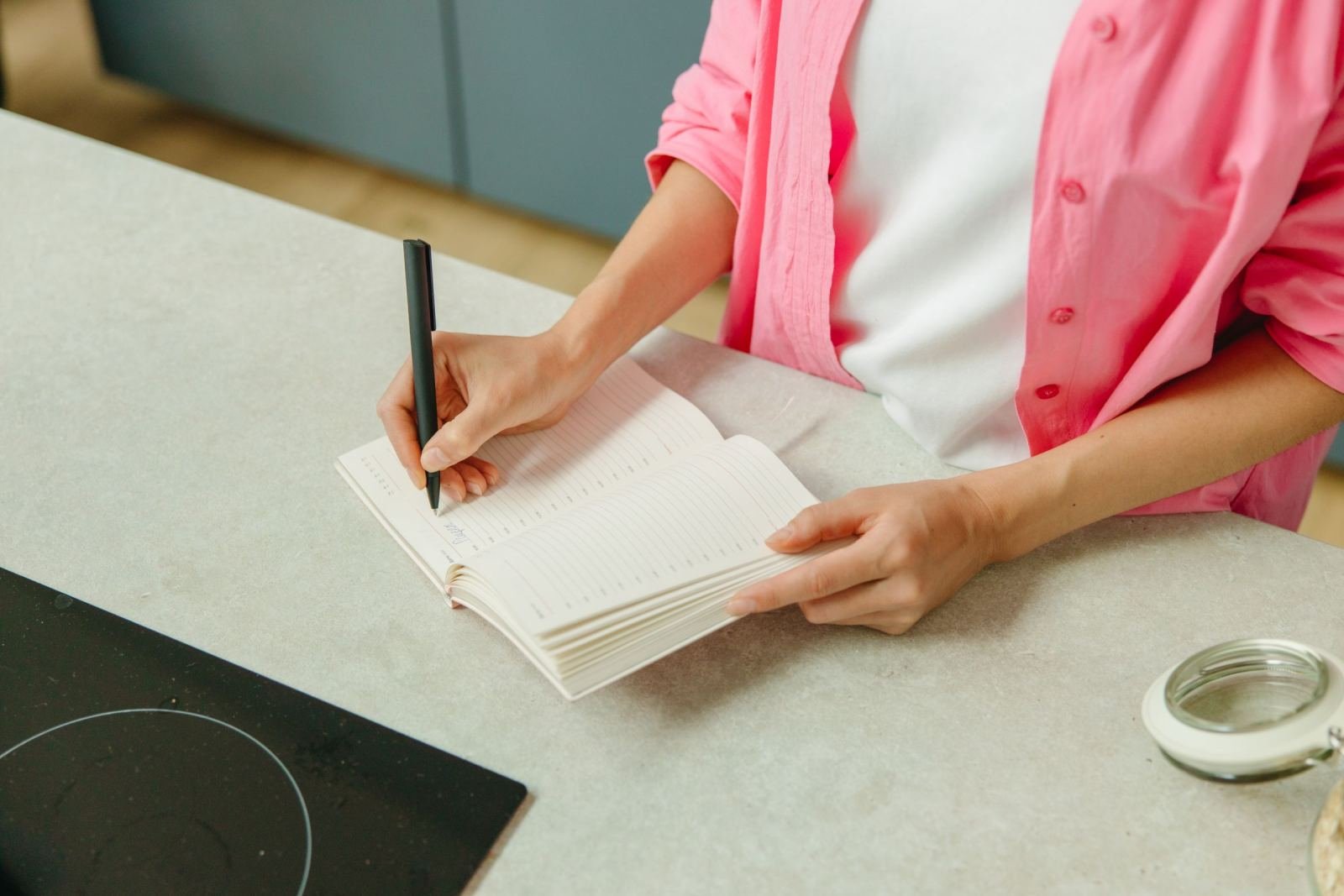 A person in a pink shirt writes in a notebook at a kitchen counter