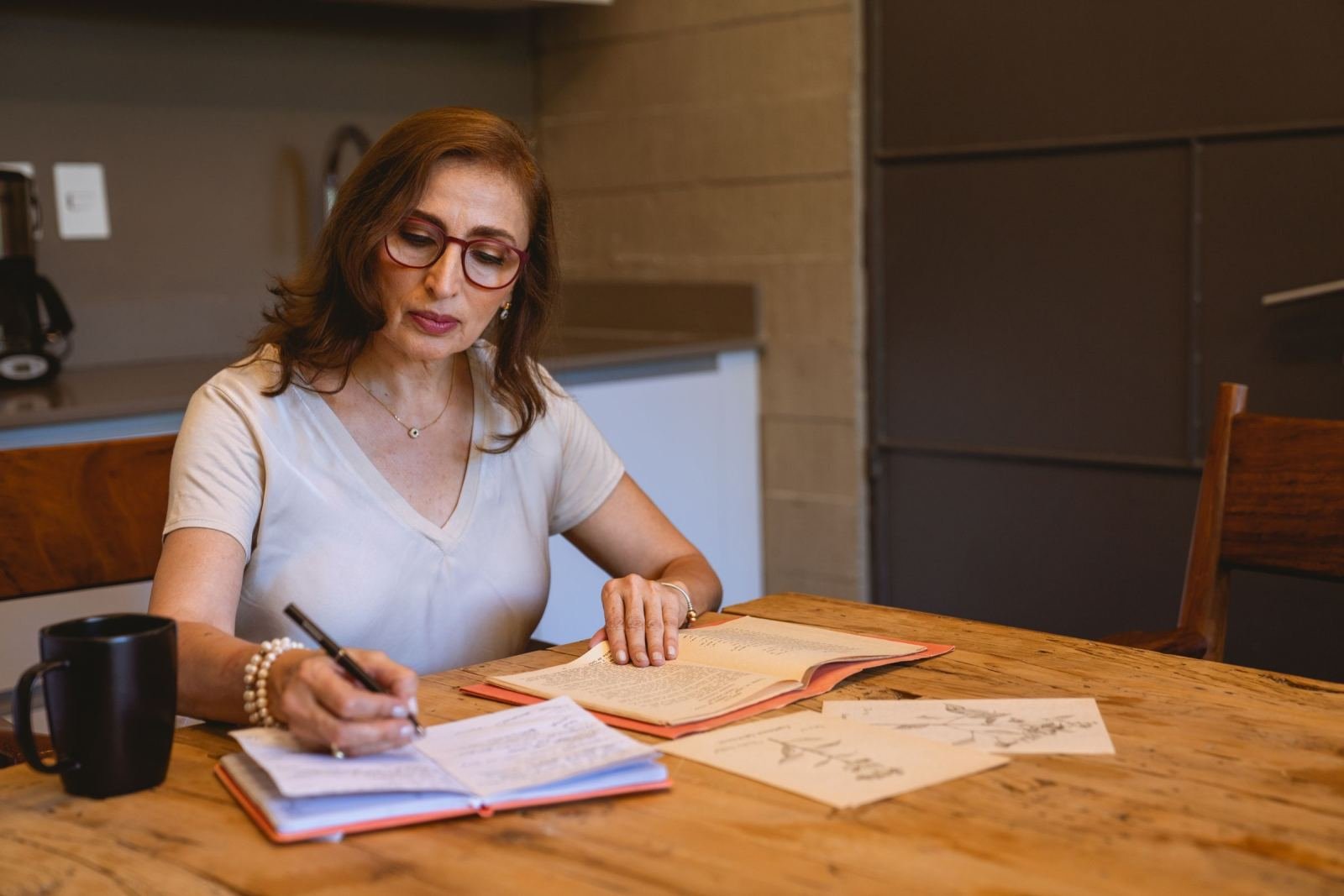 A woman with glasses writes in a notebook at a wooden table