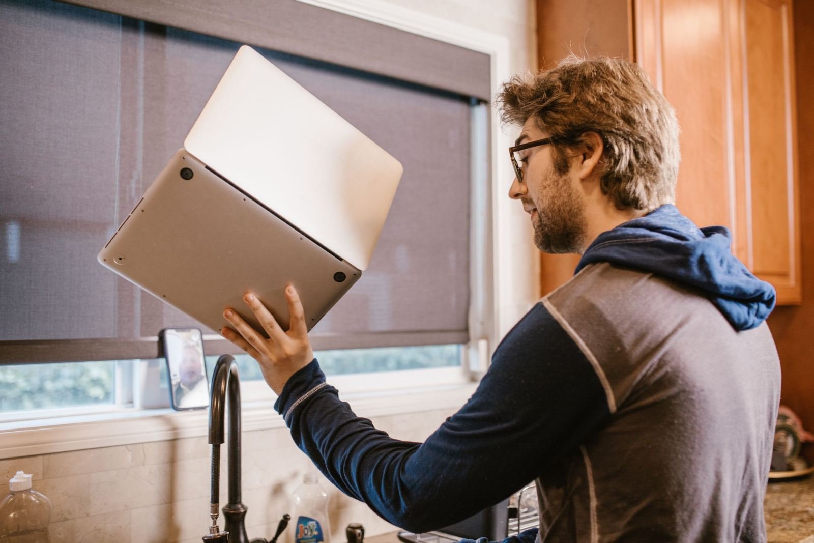 A man in a hoodie holds a laptop upside down in a kitchen