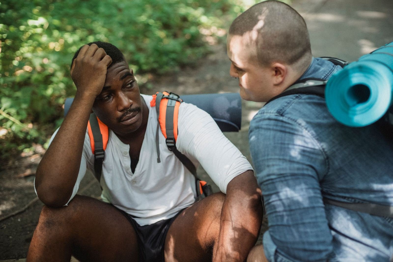 Two men with backpacks sit on a path, one with his hand on his head
