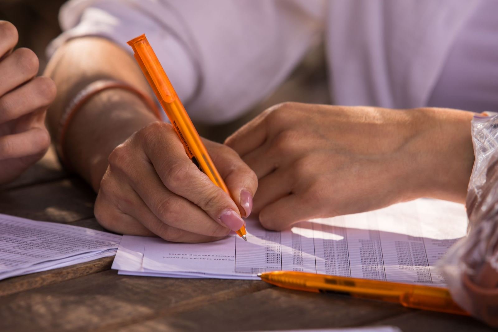 A person writes on a document with an orange pen at a wooden table