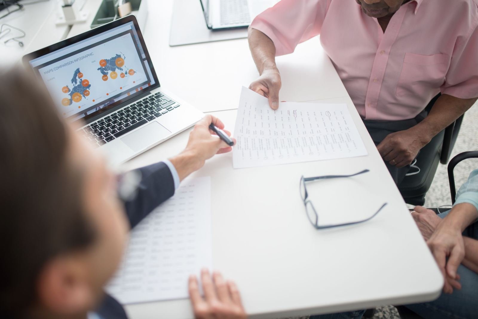 A pair of people look over a paper printout side by side at a bright table. A laptop sits nearby.