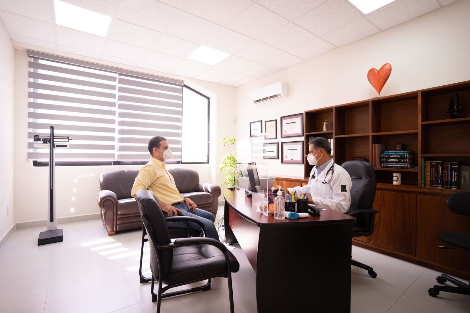 A masked doctor in a white coat chats with a patient in a sunny room.