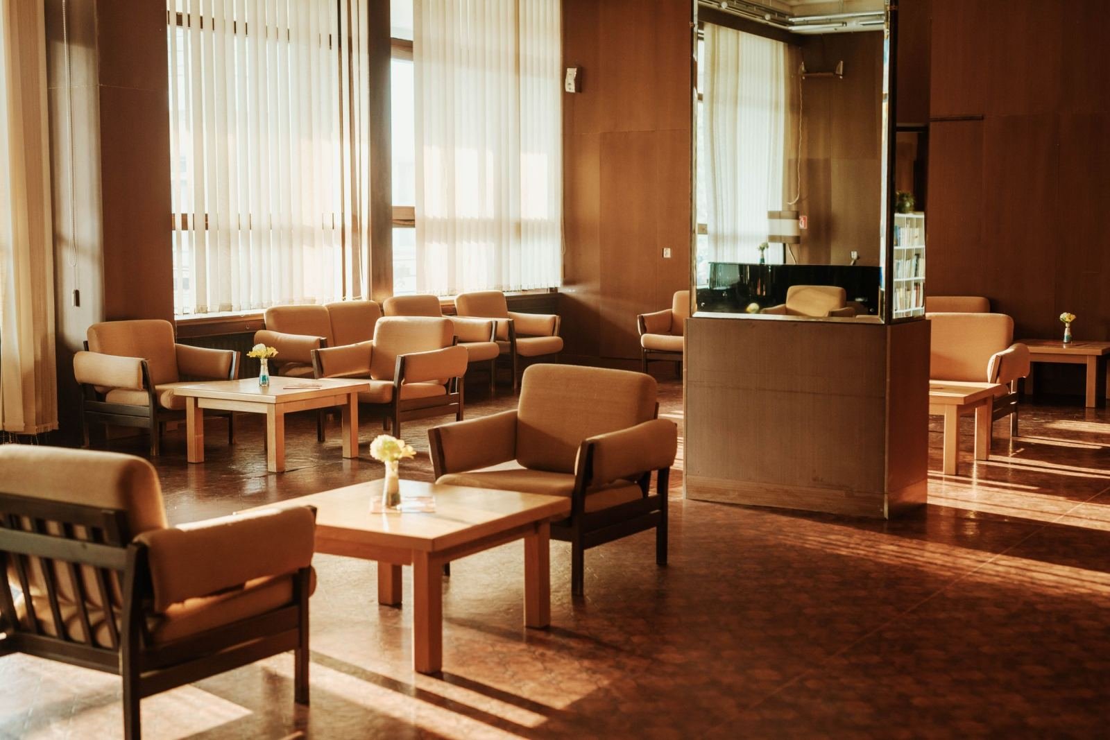 A bright common room with tan armchairs and wooden tables by large windows