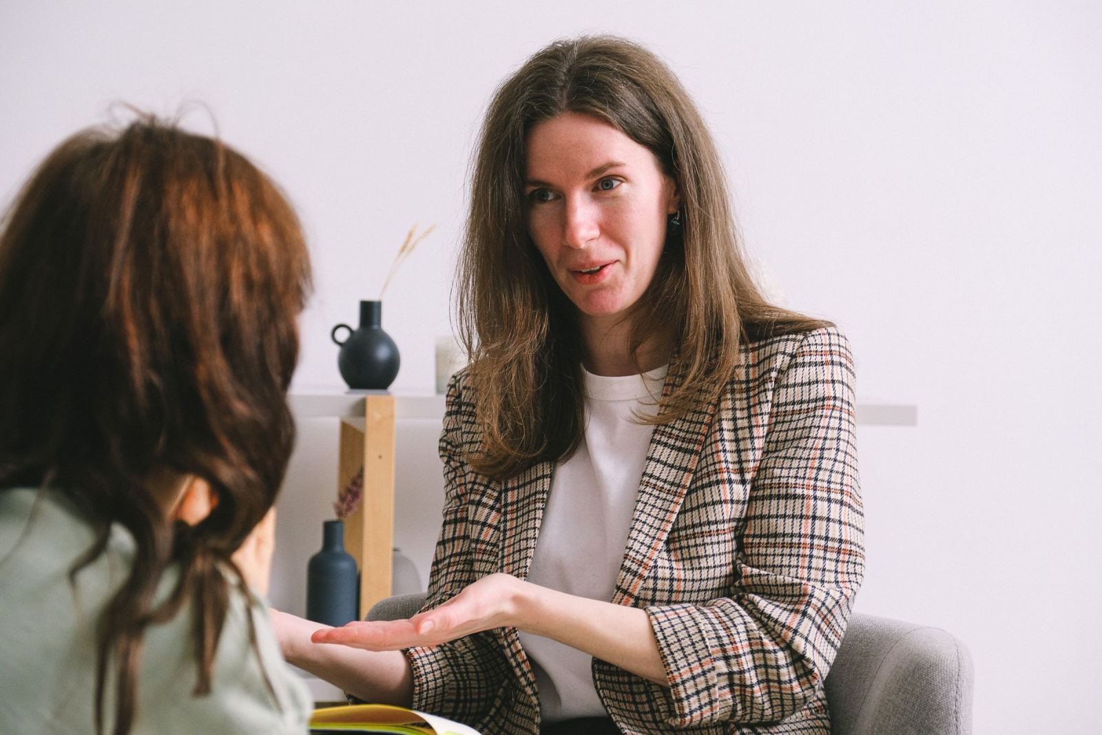 A woman in a plaid blazer speaks and gestures toward another person