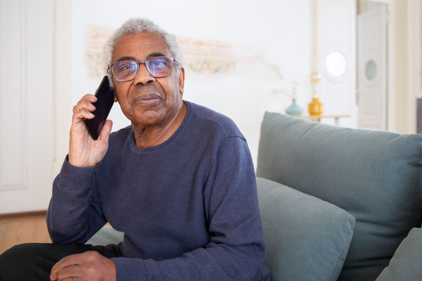 A man with older features and glasses sits on a couch. He uses a smartphone for a call.