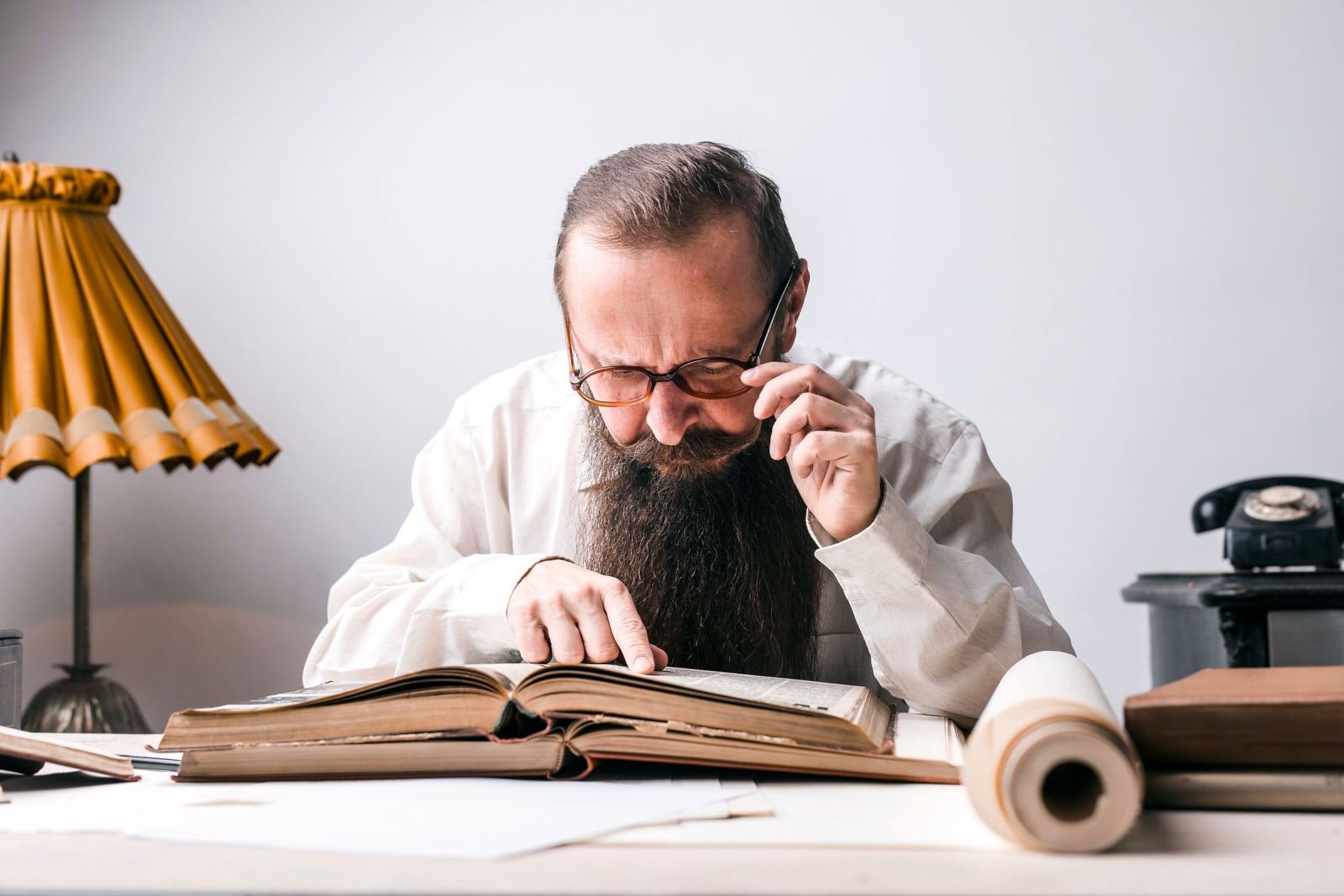 A bearded man wearing glasses reads a big open book while sitting at a desk.