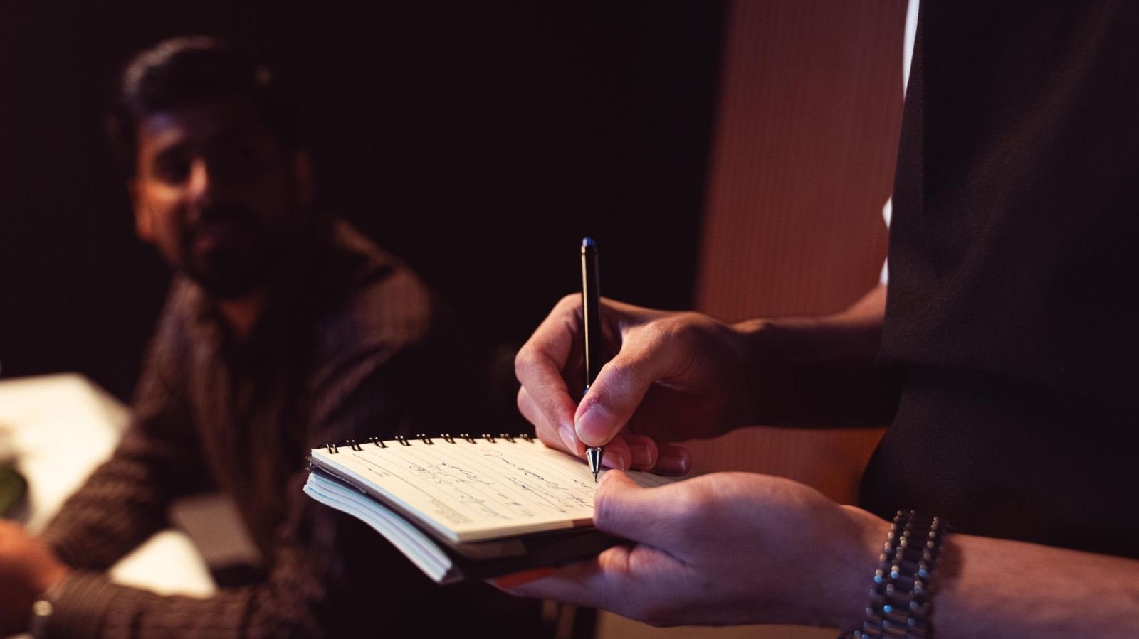 A person writes in a spiral notebook while another man sits in the background
