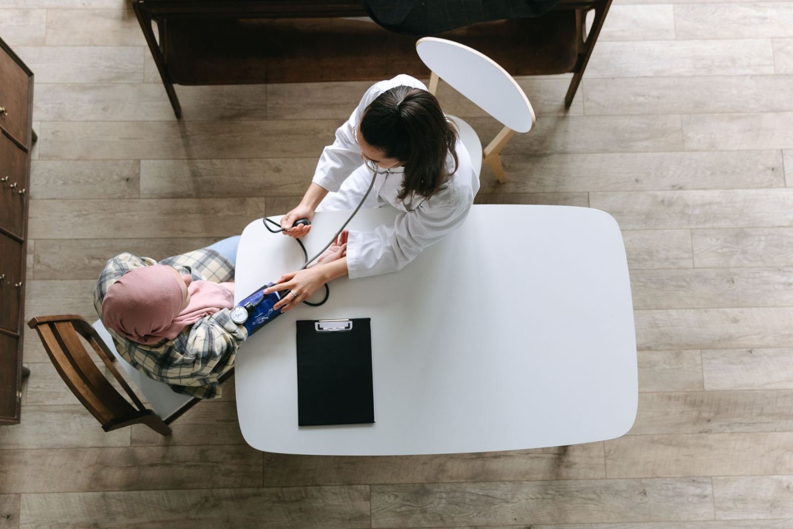A clinician wearing a white coat checks a patient's blood pressure at a white table.