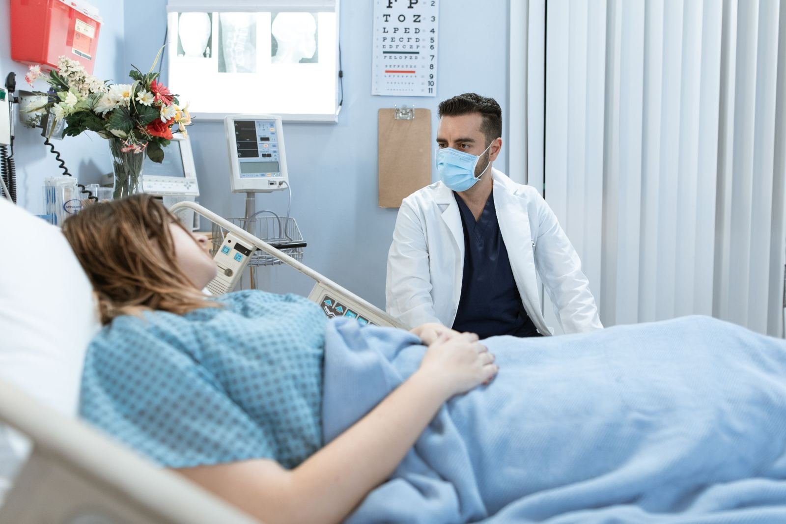 A doctor in a mask stands beside a patient lying in a hospital bed