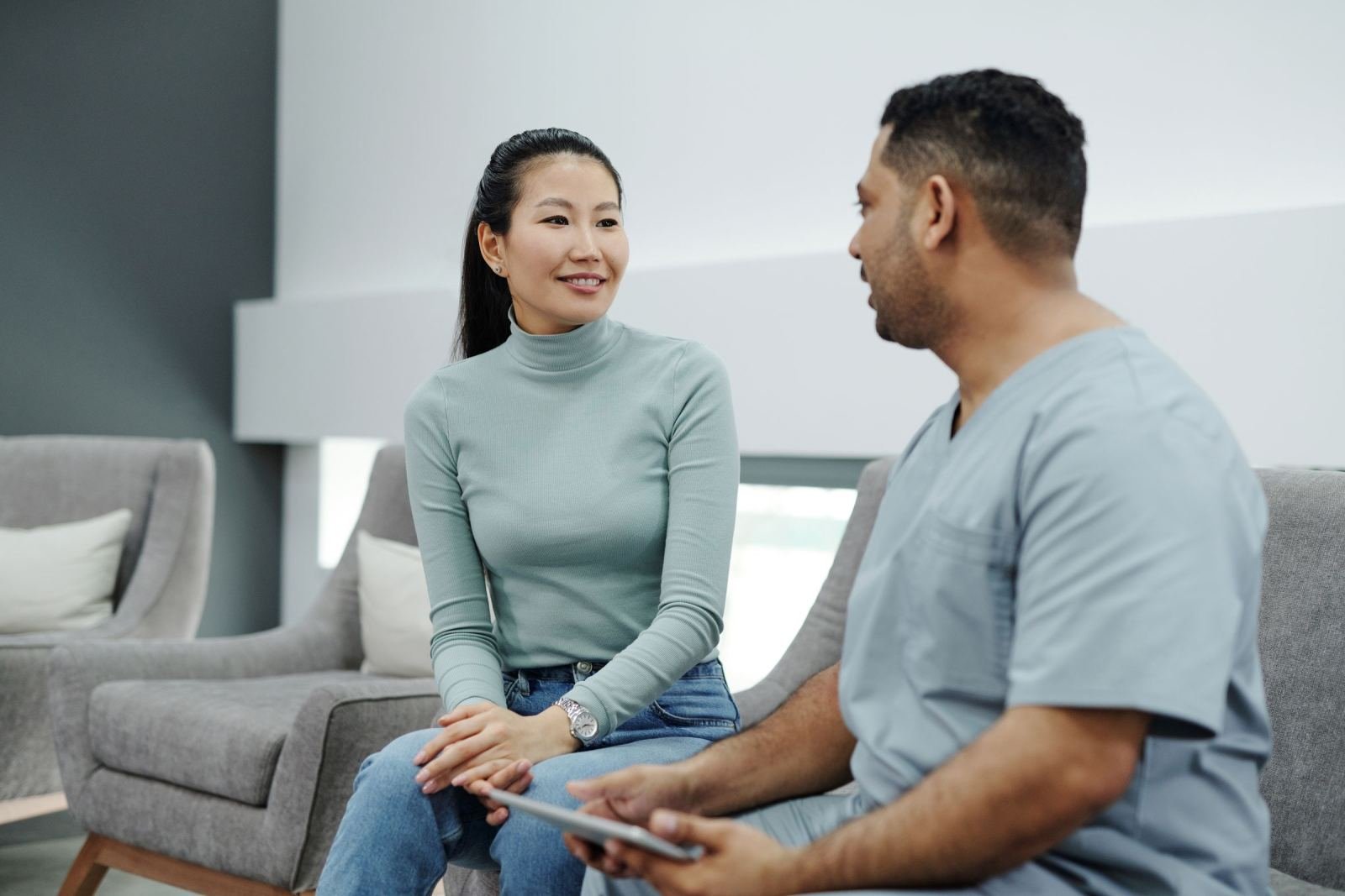 A healthcare provider in blue scrubs talks with a woman wearing a green shirt.