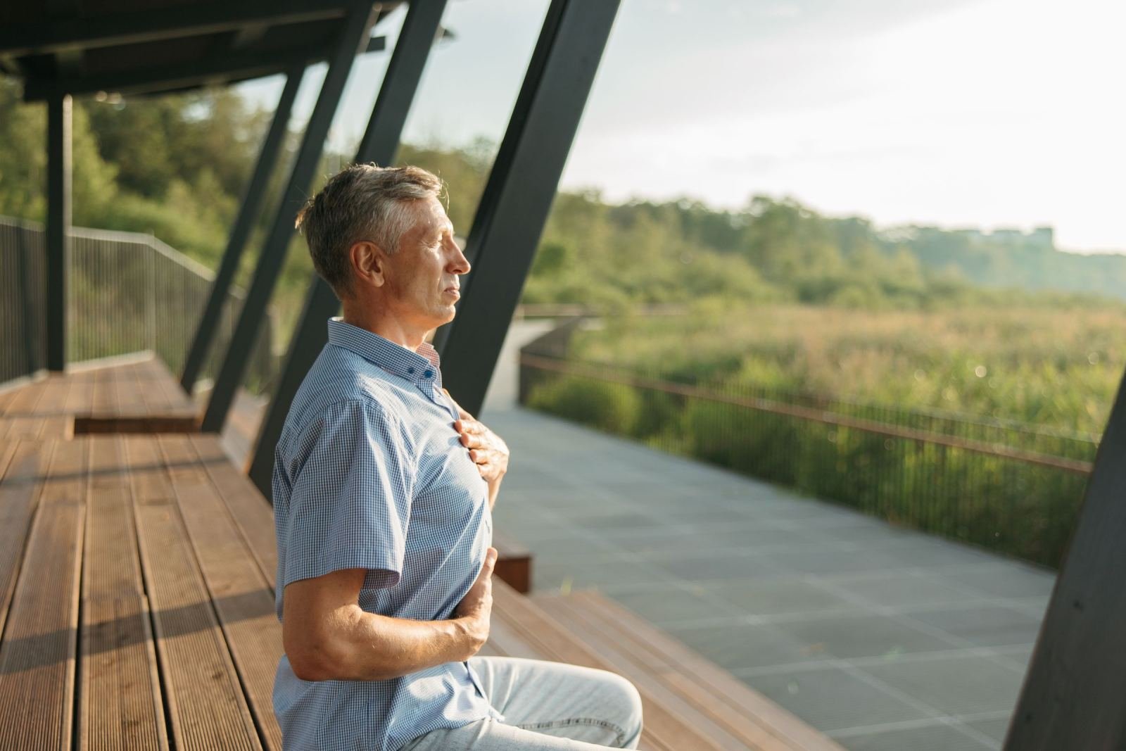 A man sits on a wooden bench with his eyes closed and a hand on his chest