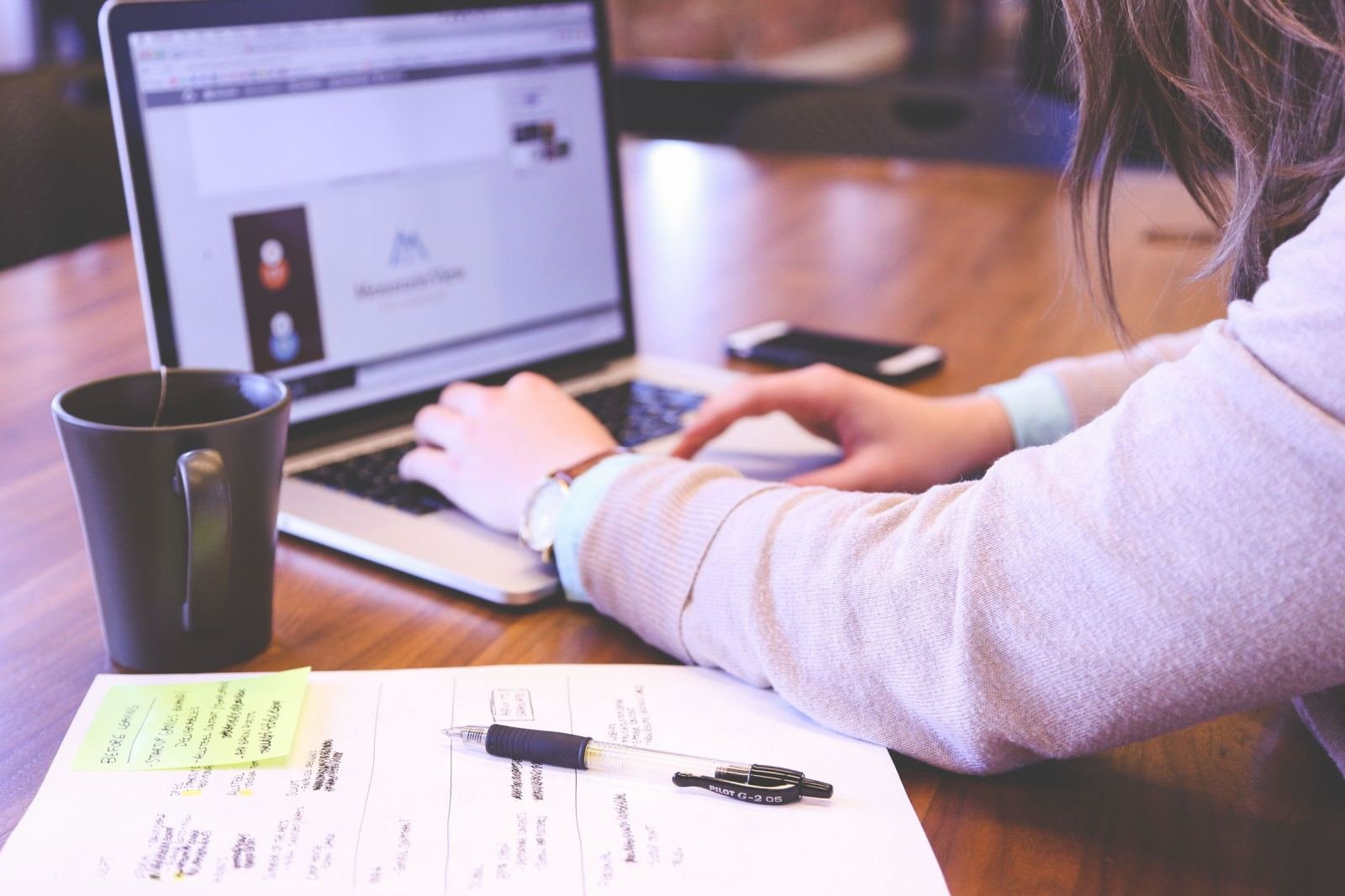 A person sits at a desk using a laptop, with a notebook and a mug nearby.