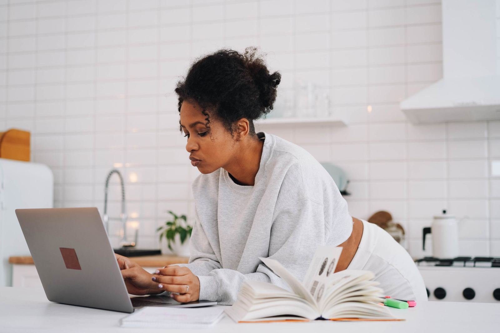 A woman looks at a laptop and an open notebook at a white kitchen counter