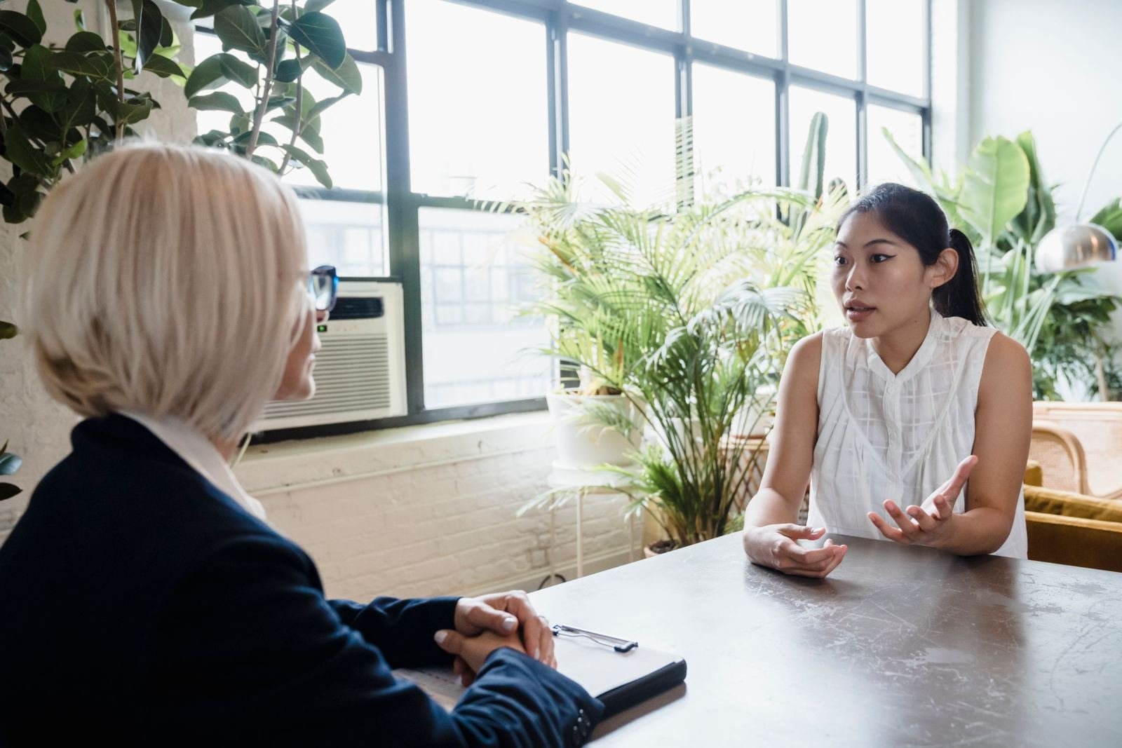 Two women sit across from each other at a table. The office is bright, with big windows and plants.