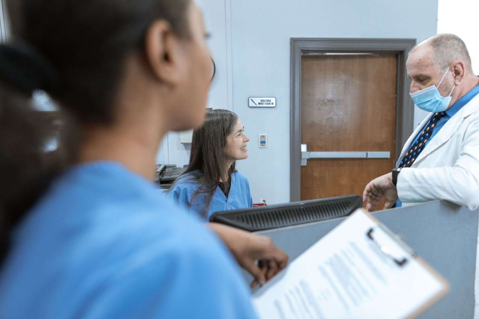 A man in a white lab coat and mask speaks with two staff members in blue scrubs