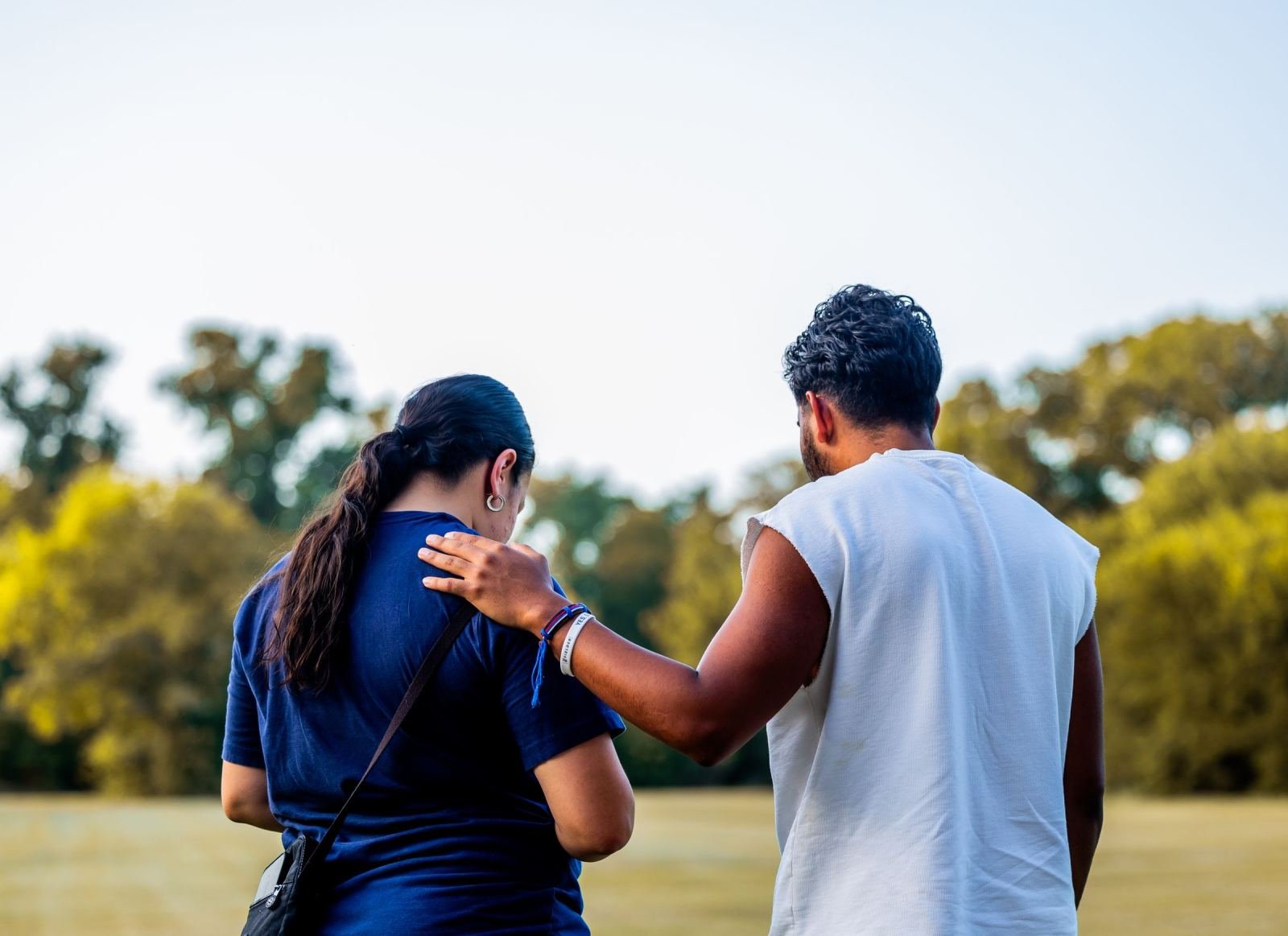 Two people stroll through a park, with one resting a comforting hand on the other's shoulder.