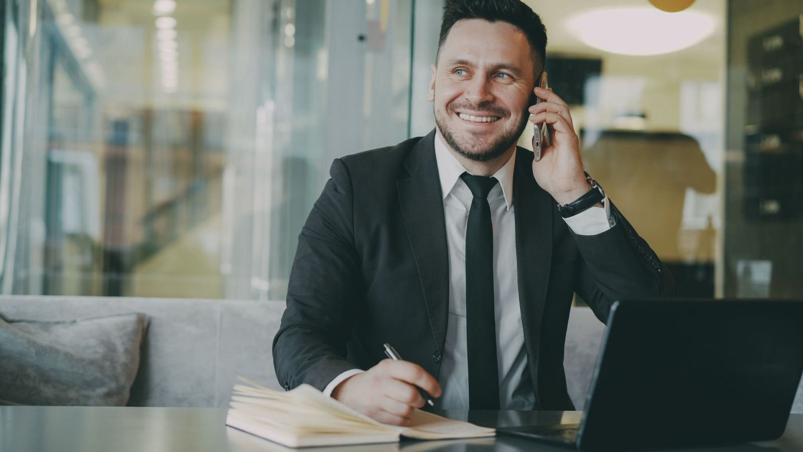 A man in a suit smiles while talking on a phone at a desk with a laptop