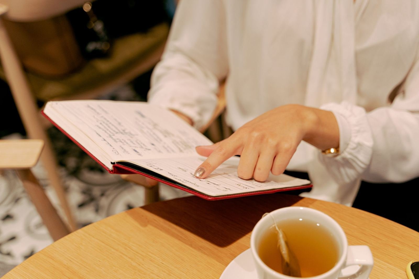 A woman in a white blouse points to notes in a red notebook. A cup of tea sits nearby.