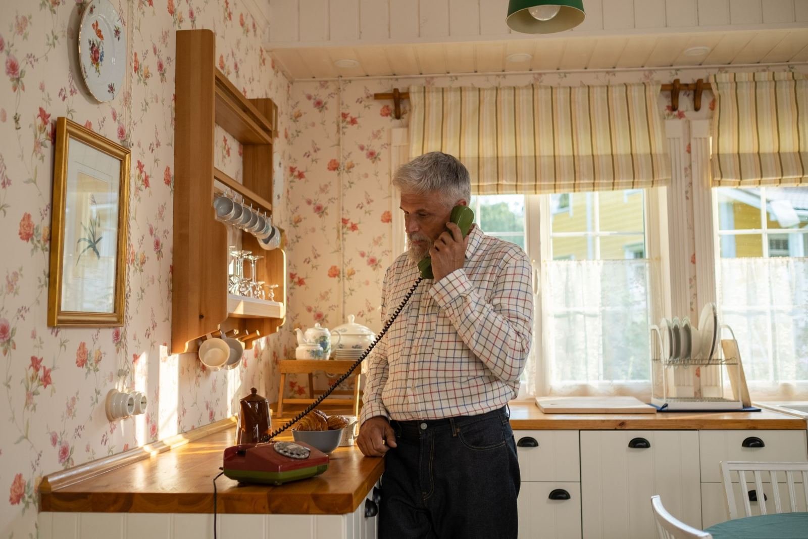 A man wearing a plaid shirt speaks on a green corded telephone while standing in a kitchen.