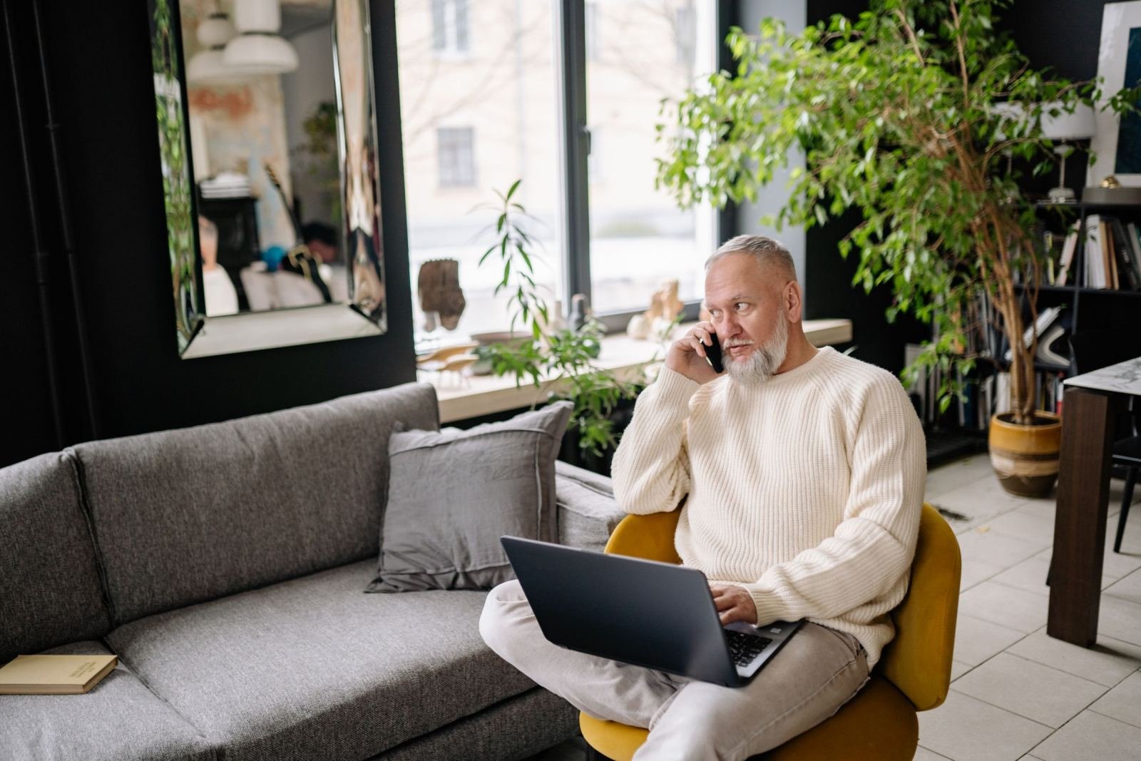 A man wearing a white sweater uses a phone and laptop in a living room.