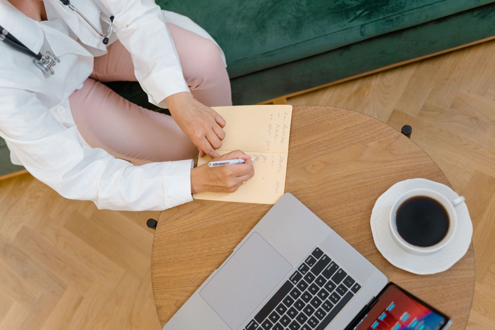 A healthcare provider in a white coat writes in a notebook next to a laptop and coffee