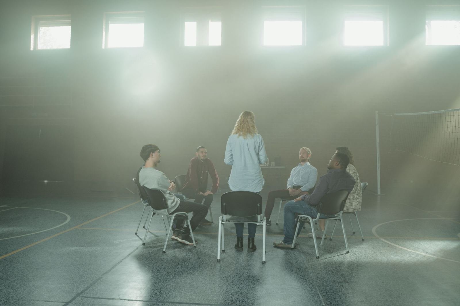 A woman stands before a circle of chairs where adults sit in a bright room.