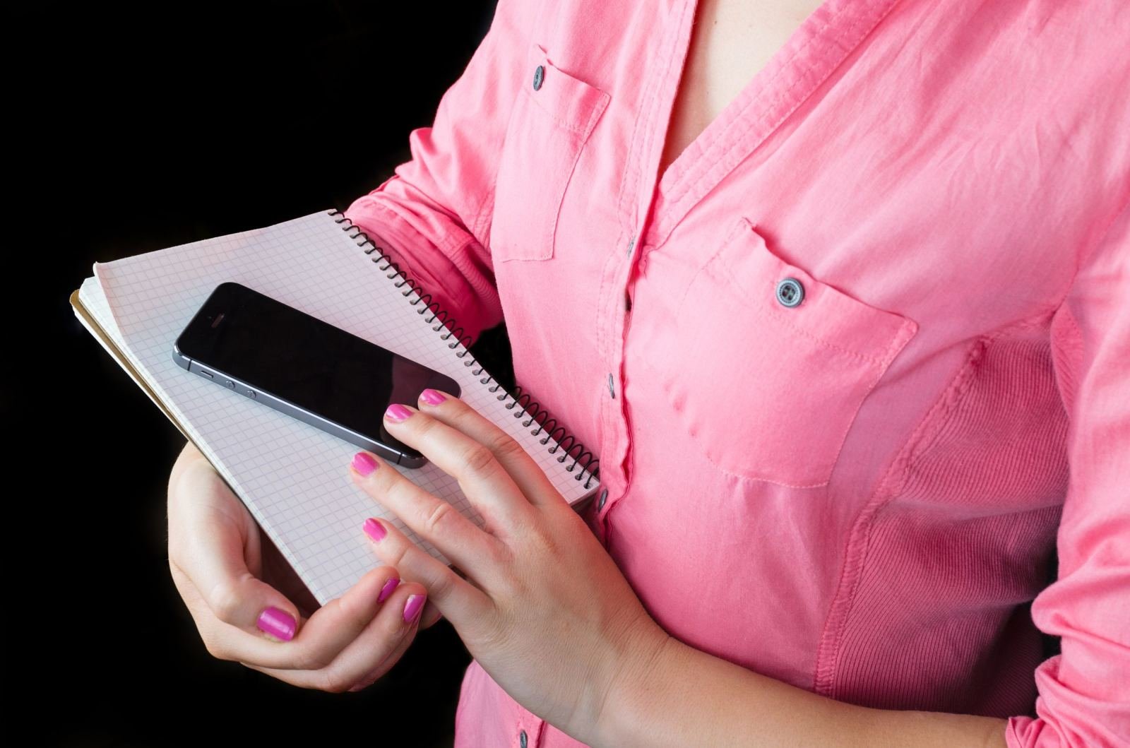 A person in a pink shirt holds a smartphone and a spiral notebook