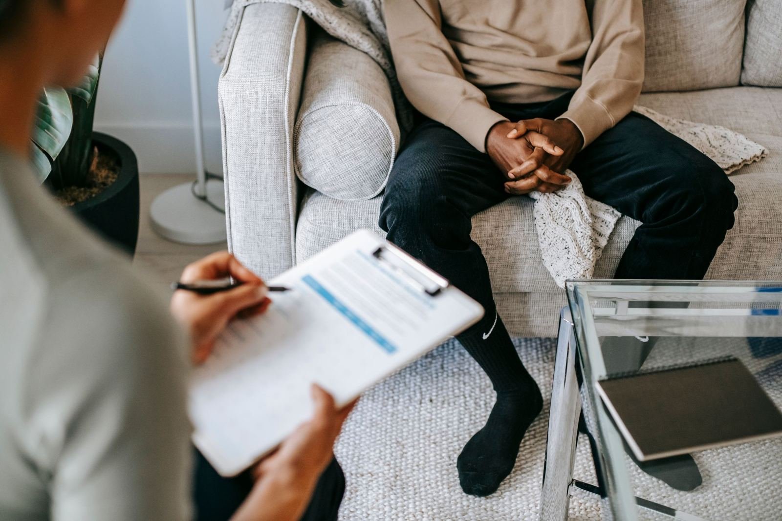 A man sits on a sofa while a therapist holds a clipboard across from him