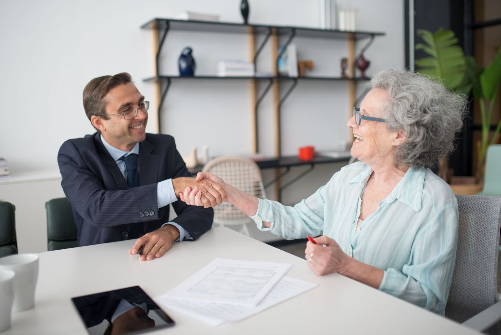 A man in a suit and an older woman shake hands across a table with papers