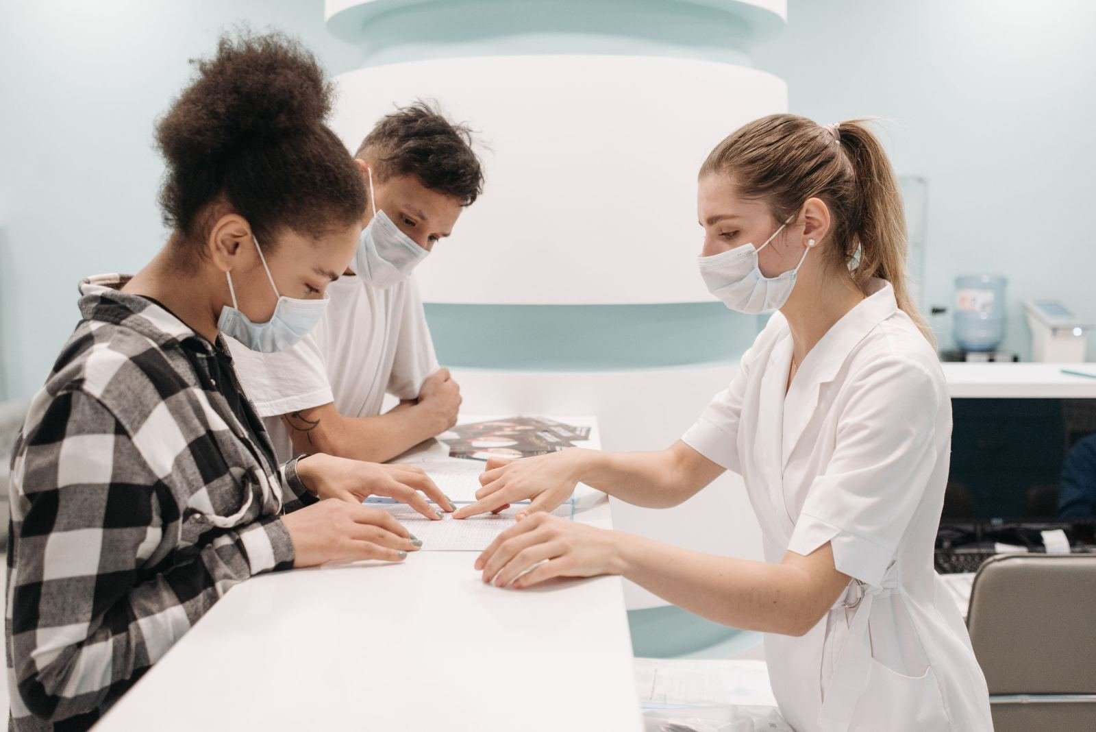 A masked clinician in a white coat goes over papers with two people at a counter.