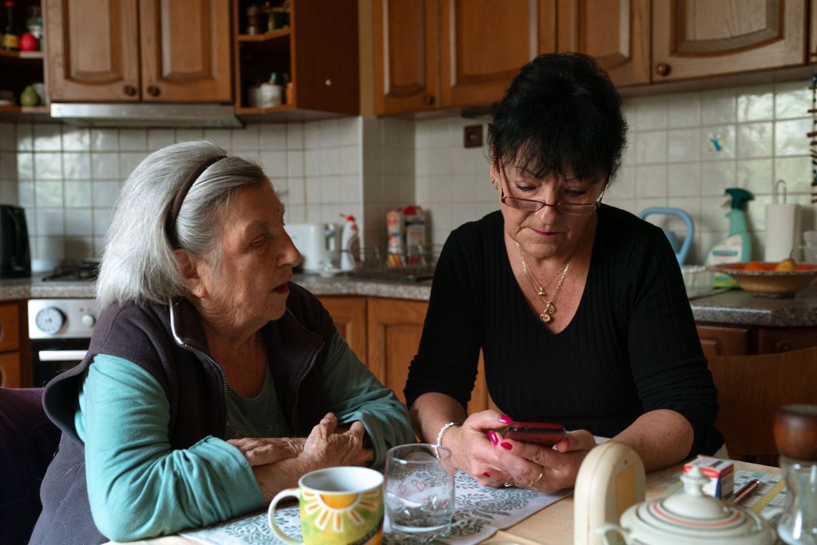 Two women sit at a kitchen table while one looks at a smartphone