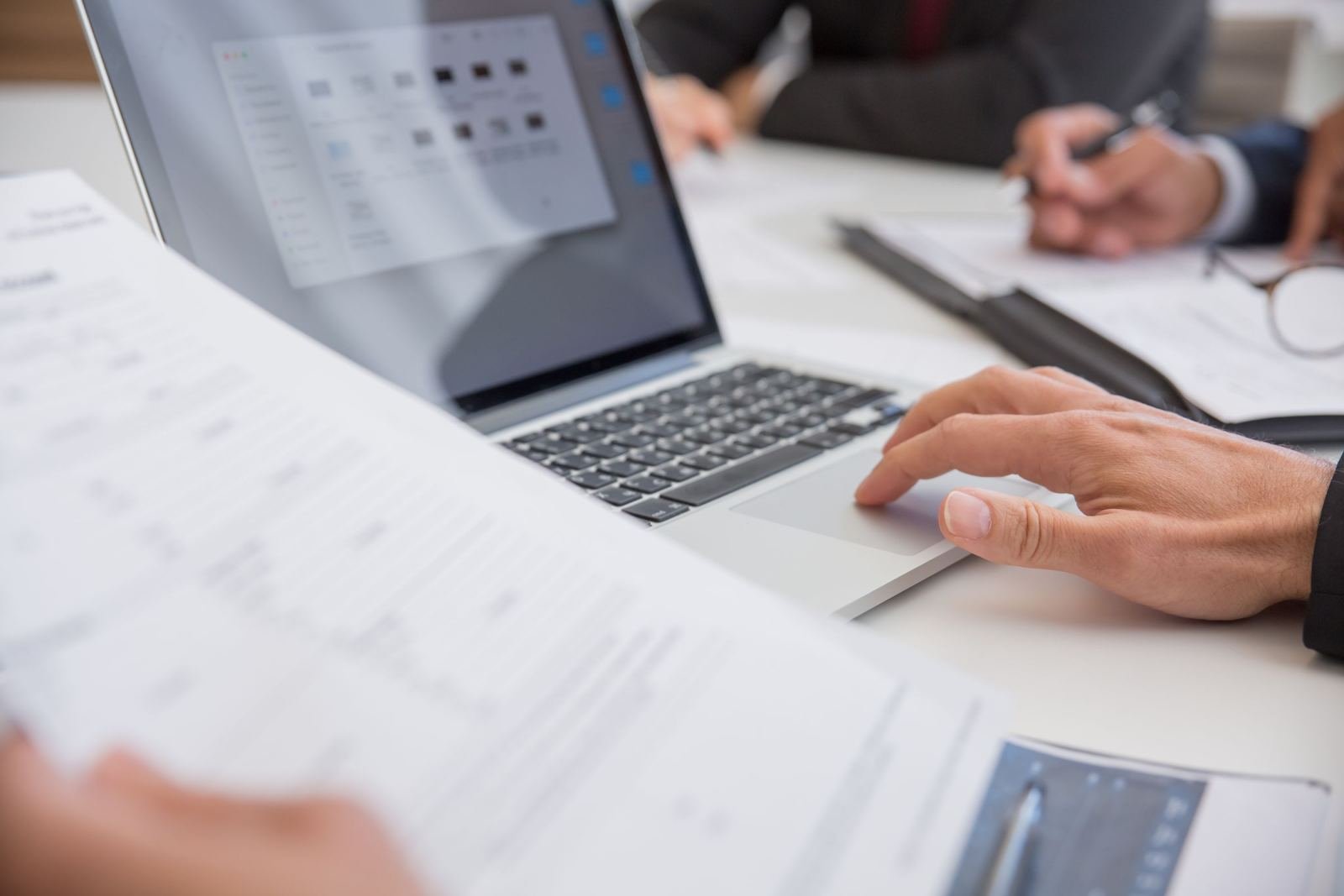 Hands using a laptop and reviewing printed documents on a white desk