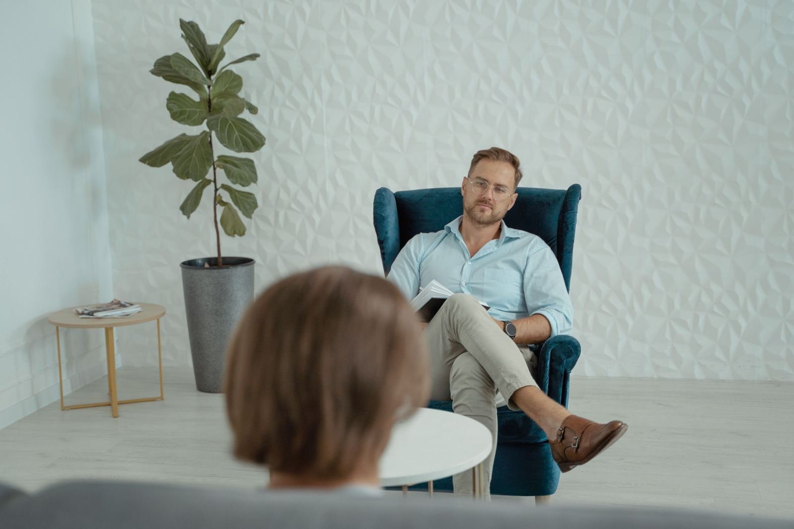 A man in a blue armchair holds a notebook while talking to a person seen from behind