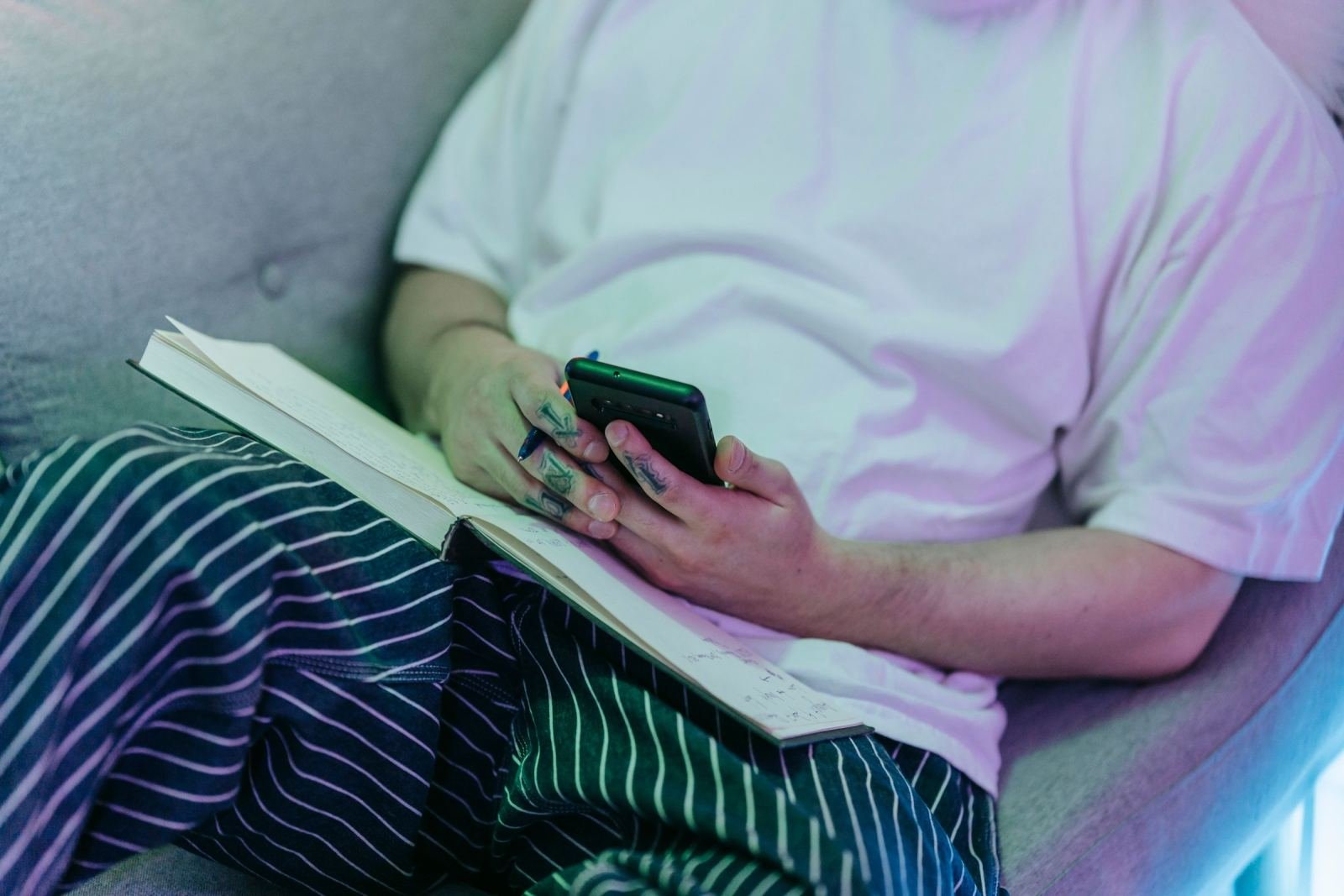 A person in pajamas sits on a sofa using a smartphone with an open notebook on their lap