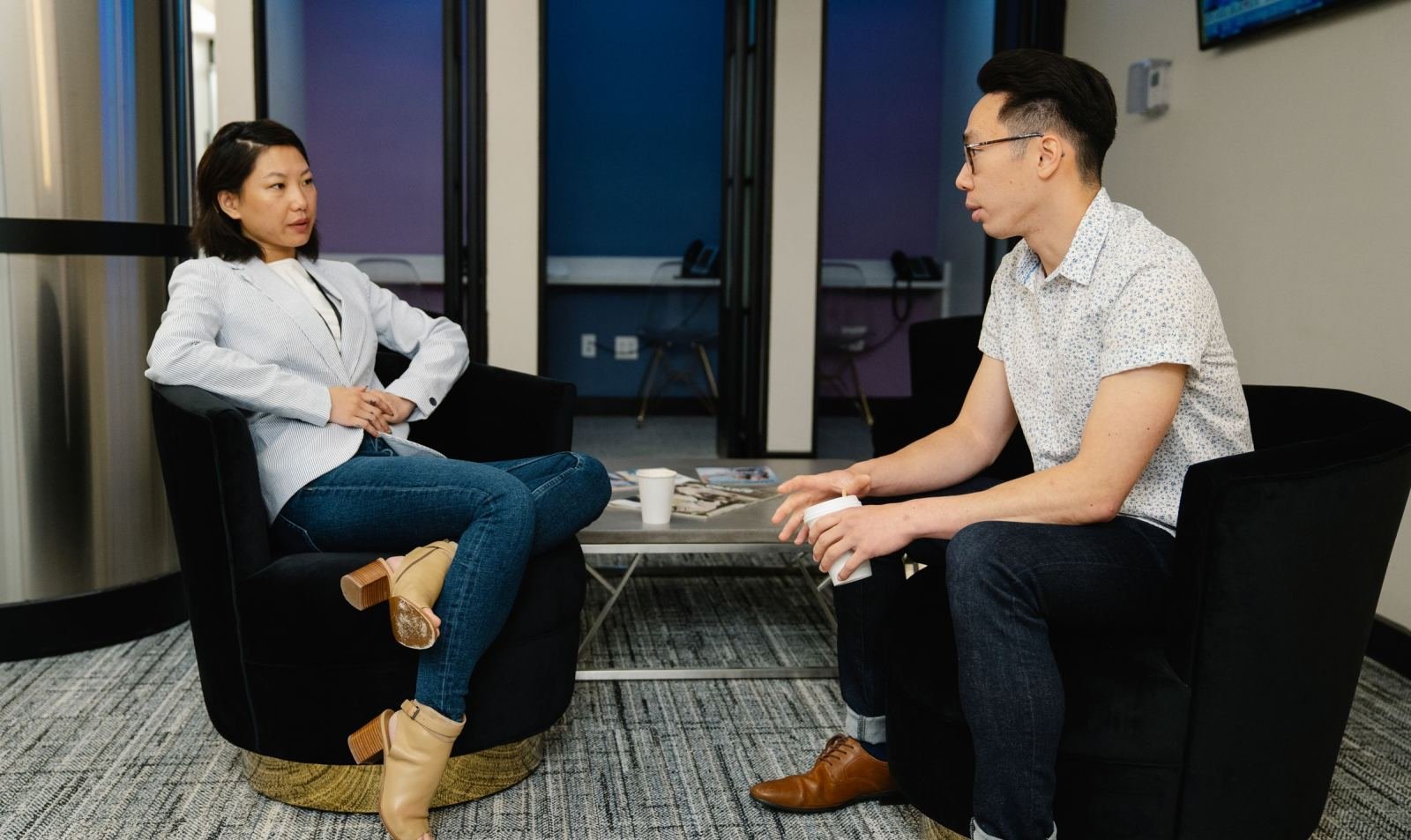 A man and woman sit in black armchairs having a conversation in a modern office