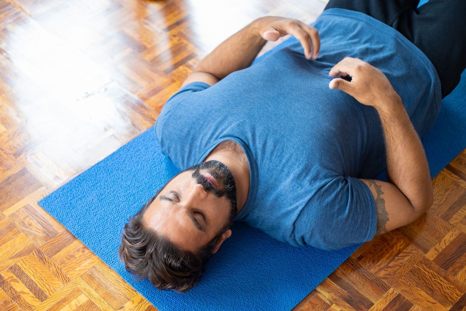 A man with his eyes closed lies on a blue yoga mat on a wooden floor